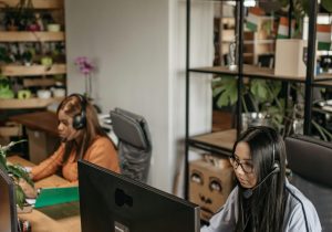Two women in headsets working diligently in a modern office space with green plants and industrial design elements.